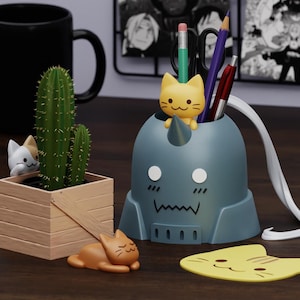 May include: A blue and gray desk organizer shaped like a cartoon character with a cat head and a horn. The organizer has a cat head on the top and a cat head on the bottom. There are two pencils, a pen, and a yellow cat figurine inside the organizer. There is a yellow cat figurine on the table next to the organizer.