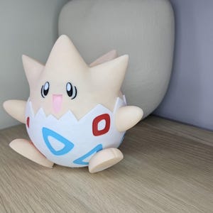 May include: A Togepi figurine, a Pok&eacute;mon character, is displayed on a wooden surface. The figure is primarily cream-colored with a white eggshell design, featuring red and blue accents. It has small arms and legs, and a smiling expression.