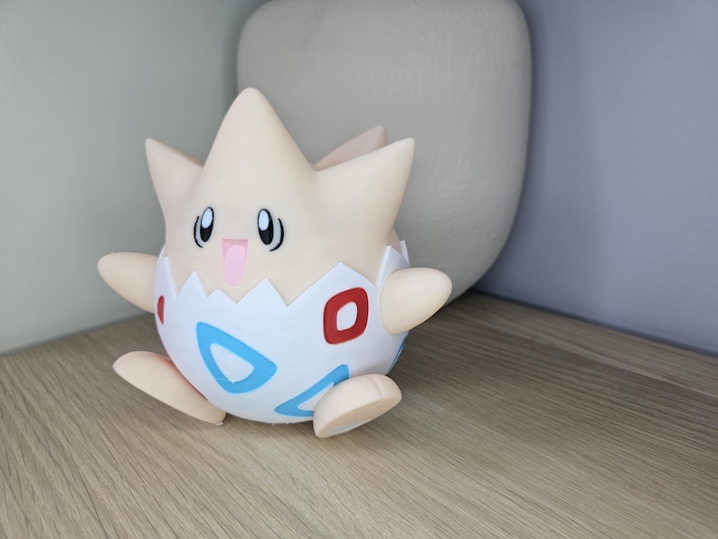 May include: A Togepi figurine, a Pok&eacute;mon character, is displayed on a wooden surface. The figure is primarily cream-colored with a white eggshell design, featuring red and blue accents. It has small arms and legs, and a smiling expression.