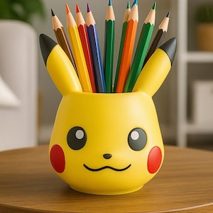 Puede incluir: Un portalápices amarillo con forma de Pikachu lleno de lápices de colores. El portalápices tiene orejas, ojos y boca sonriente negros, con círculos rojos en las mejillas. Los lápices son de varios colores, incluyendo marrón, naranja, rojo, morado, azul y verde.