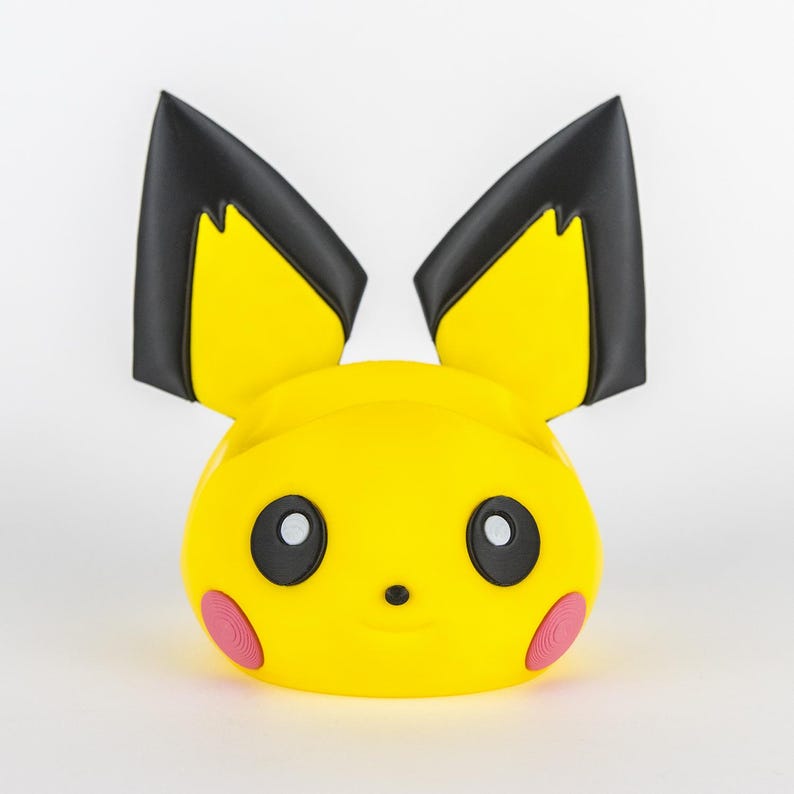 Pu&ograve; includere: Un supporto per telefono giallo a forma di Pikachu, con orecchie con punte nere e accenti rosa sulle guance. Il supporto ha una faccia sorridente con occhi neri e un piccolo naso. Il design &egrave; una rappresentazione a cartoni animati del personaggio popolare.