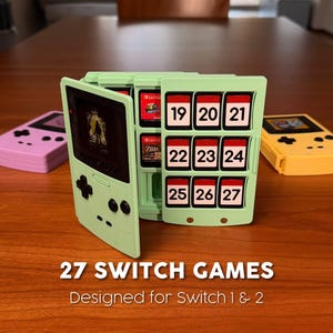 May include: A light green game cartridge holder shaped like a retro handheld console, holding 27 Switch games. The holder has numbered slots and the text "27 SWITCH GAMES" and "Designed for Switch 1 & 2". Two other game consoles are on the table.