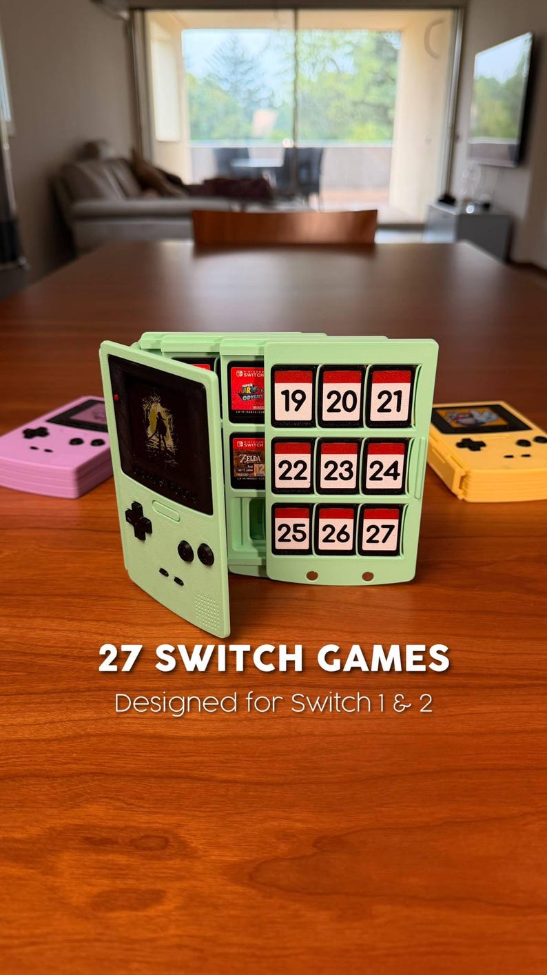 May include: A light green game cartridge holder shaped like a retro handheld console, holding 27 Switch games. The holder has numbered slots and the text "27 SWITCH GAMES" and "Designed for Switch 1 & 2". Two other game consoles are on the table.