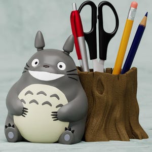 May include: A gray and white Totoro character desk organizer with a brown tree trunk base. The organizer holds a red pen, a silver pen, a pair of black scissors, a yellow pencil, and a blue pencil.