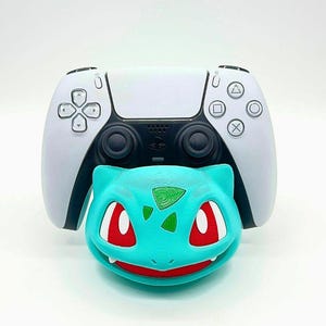 Puede incluir: Un mando de PlayStation 5 blanco y negro con un accesorio en forma de Bulbasaur turquesa. El accesorio Bulbasaur tiene ojos rojos y boca roja. El mando tiene una cruceta, botones y dos joysticks.