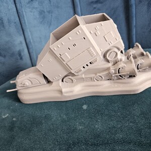 May include: A gray 3D printed model of a Star Wars AT-AT walker, with the walker tipped over on its side. The model is on a base and has detailed features.