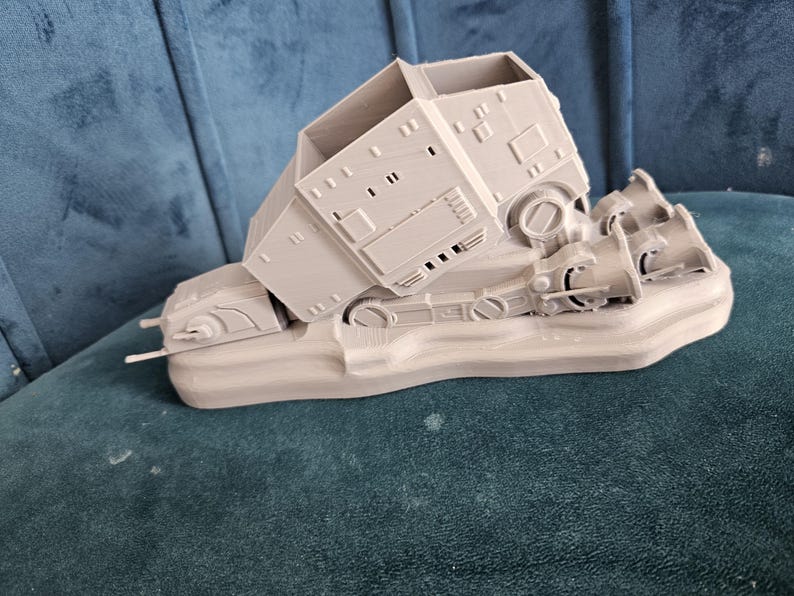 May include: A gray 3D printed model of a Star Wars AT-AT walker, with the walker tipped over on its side. The model is on a base and has detailed features.
