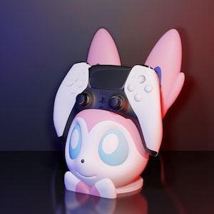 May include: A pink and white character-themed controller stand with a black and white game controller resting on top. The stand has large pink ears and blue eyes. The background is a gradient of red and blue.