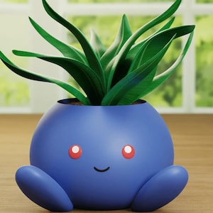 May include: A blue ceramic planter shaped like a cartoon character with red eyes and a smile. A green plant is growing out of the top of the planter.