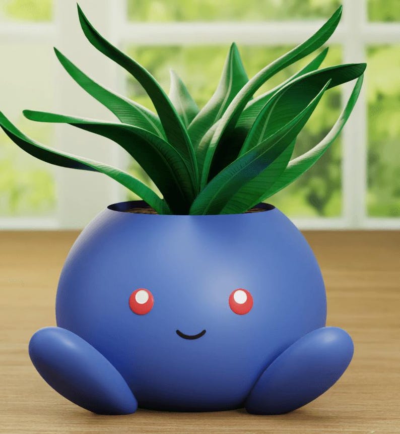 May include: A blue ceramic planter shaped like a cartoon character with red eyes and a smile. A green plant is growing out of the top of the planter.