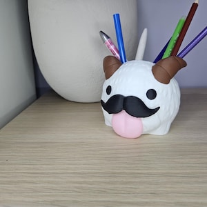 May include: White plush pen holder shaped like a cartoon animal with a black mustache and pink tongue. The animal has brown ears and a white fluffy body.
