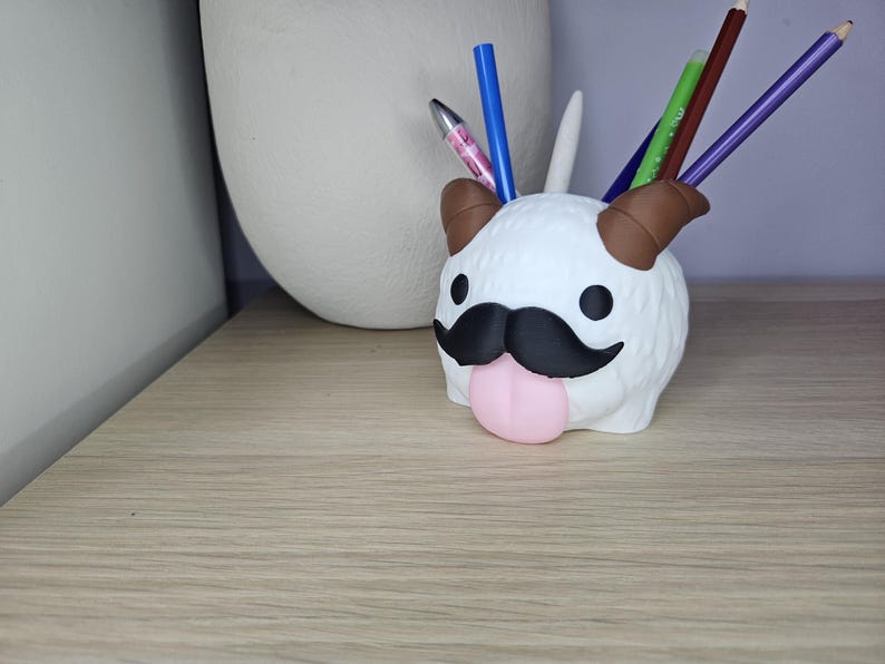 May include: White plush pen holder shaped like a cartoon animal with a black mustache and pink tongue. The animal has brown ears and a white fluffy body.