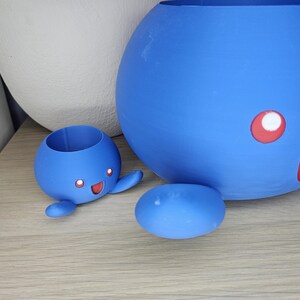 May include: Two blue ceramic planters shaped like cartoon characters. The larger planter is round and has a red mouth and white eye. The smaller planter is shaped like a small creature with arms and legs and has a red mouth and two white eyes.