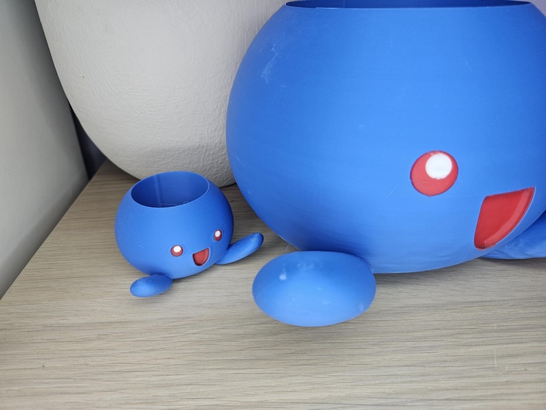 May include: Two blue ceramic planters shaped like cartoon characters. The larger planter is round and has a red mouth and white eye. The smaller planter is shaped like a small creature with arms and legs and has a red mouth and two white eyes.