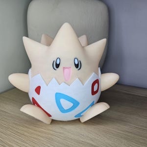 May include: A Togepi figurine, a Pok&eacute;mon character, is shown. The figure is primarily cream-colored with a star-shaped head and small arms and feet. The lower half is white with red and blue accents. The figurine is sitting on a wooden surface.