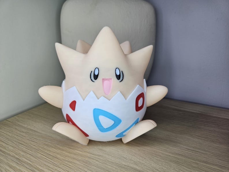 May include: A Togepi figurine, a Pok&eacute;mon character, is shown. The figure is primarily cream-colored with a star-shaped head and small arms and feet. The lower half is white with red and blue accents. The figurine is sitting on a wooden surface.