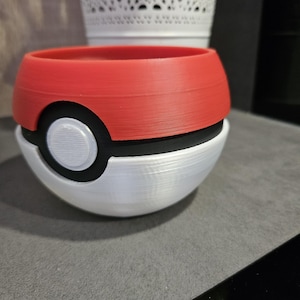 May include: A red, black, and white ceramic planter shaped like a Poke Ball. The planter has a white circle with a black outline in the center.