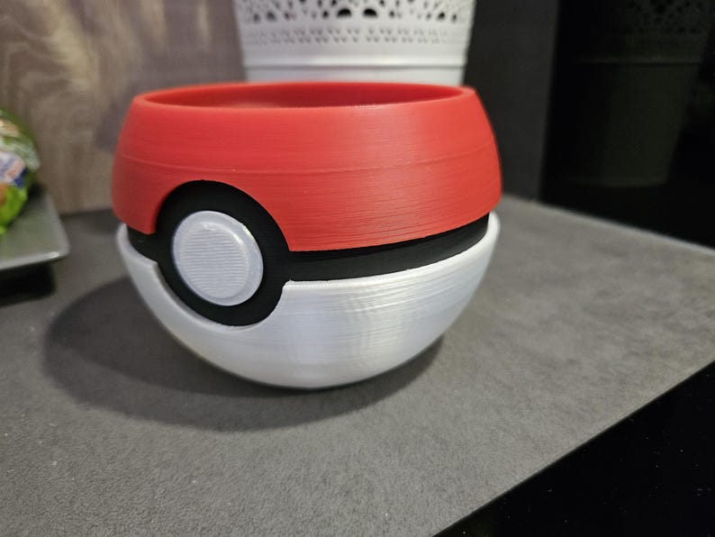 May include: A red, black, and white ceramic planter shaped like a Poke Ball. The planter has a white circle with a black outline in the center.