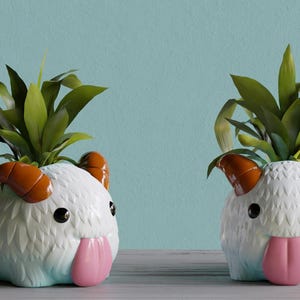May include: Two white ceramic planter pots shaped like a fluffy white creature with orange horns and a pink tongue. Each pot has a green plant growing out of the top.