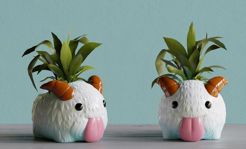 May include: Two white ceramic planter pots shaped like a fluffy white creature with orange horns and a pink tongue. Each pot has a green plant growing out of the top.