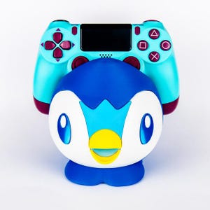 May include: A teal and maroon PlayStation 4 controller rests atop a blue, white, and yellow penguin figurine. The controller features black buttons and a black touchpad. The penguin figurine has large blue eyes and a yellow beak.