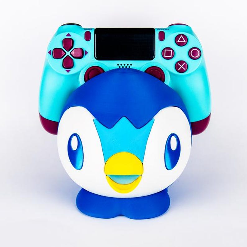 May include: A teal and maroon PlayStation 4 controller rests atop a blue, white, and yellow penguin figurine. The controller features black buttons and a black touchpad. The penguin figurine has large blue eyes and a yellow beak.