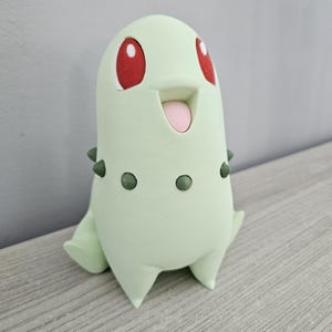 Op de afbeelding: Een lichtgroen Chikorita-beeldje met rode ogen en een roze tong. Het beeldje heeft kleine groene stekels op zijn lichaam en staat met de benen iets uit elkaar. Het beeldje staat op een lichtgrijze ondergrond.