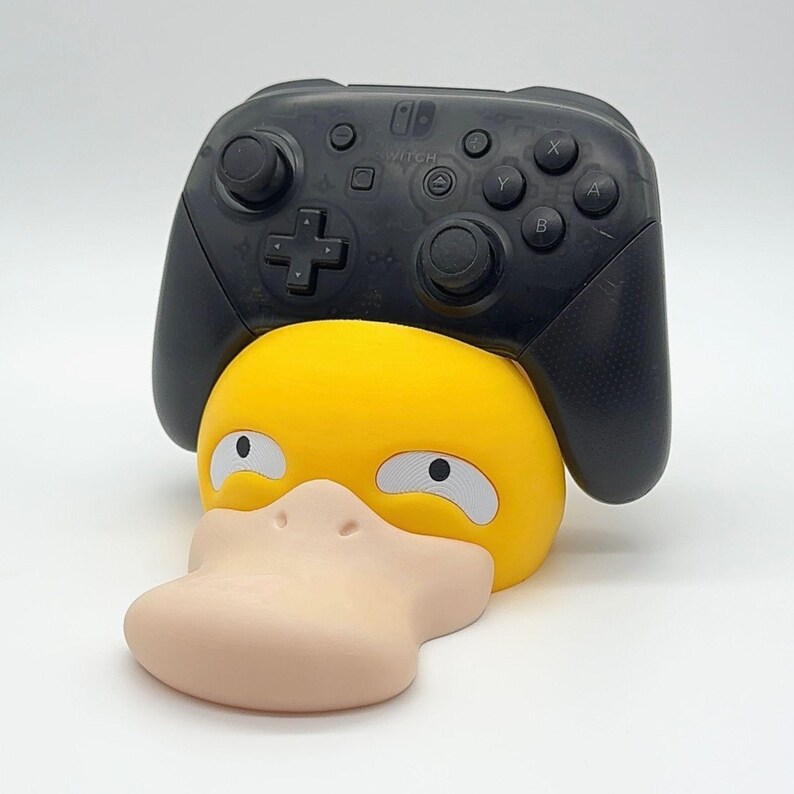 May include: A black Nintendo Switch Pro Controller rests on a yellow and tan Psyduck-shaped controller holder. The controller holder has white eyes and a tan beak. The controller has buttons and directional pads.