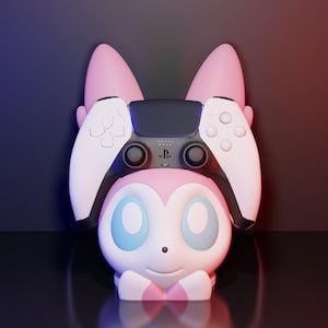 May include: A pink and white gaming controller holder shaped like a cartoon character with large ears. The holder is designed to cradle a black and white gaming controller. The background is a gradient of dark blue and red.