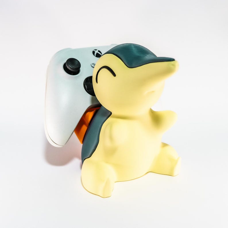 May include: A yellow and teal Cyndaquil figure holding a white Xbox controller. The figure has a smiling face and is in a sitting position. The controller has black buttons and a black joystick.