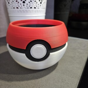 May include: A red and white Pokeball-shaped planter with a black rim and a white center circle. The planter is made of 3D printed plastic.