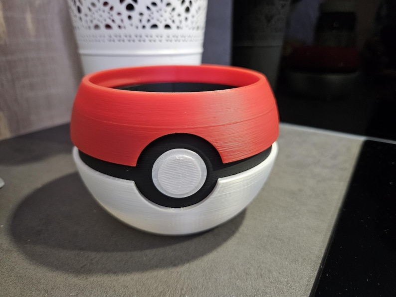 May include: A red and white Pokeball-shaped planter with a black rim and a white center circle. The planter is made of 3D printed plastic.