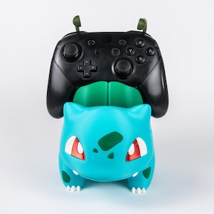 May include: A turquoise and green creature-shaped controller stand holding a black wireless game controller. The stand features a rounded body, two small ears, and red accents on the face, providing a stable base for the gaming peripheral.