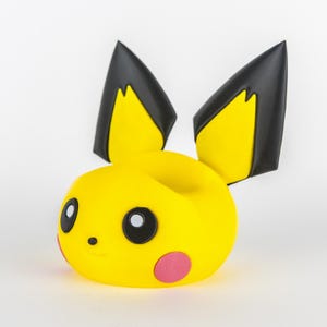 May include: A yellow Pikachu-shaped phone holder with black and yellow ears, black eyes, a small black nose, and pink cheek circles. The holder is designed to cradle a phone or other small device.