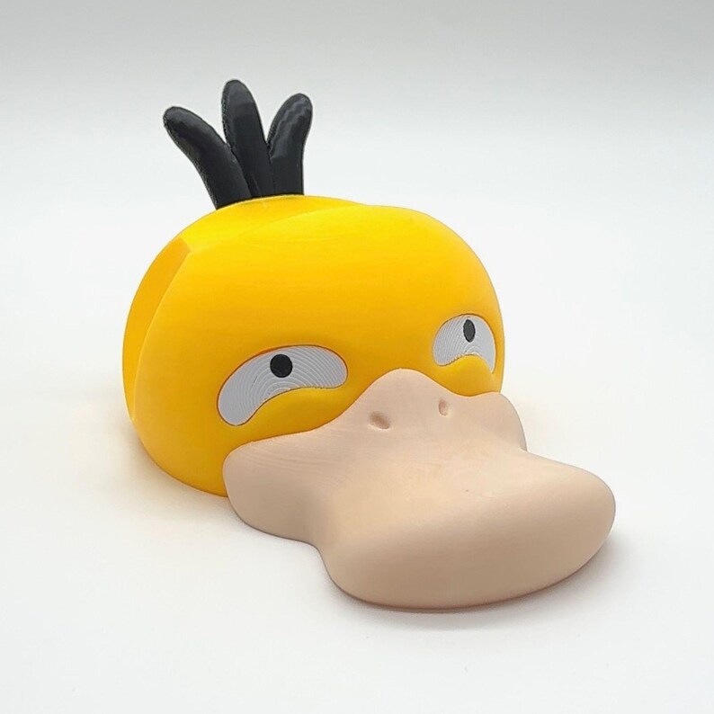 May include: A yellow, duck-shaped figurine with a large, light-tan beak and gray eyes. The figurine has a black crest on top of its head. The design is reminiscent of a popular cartoon character.