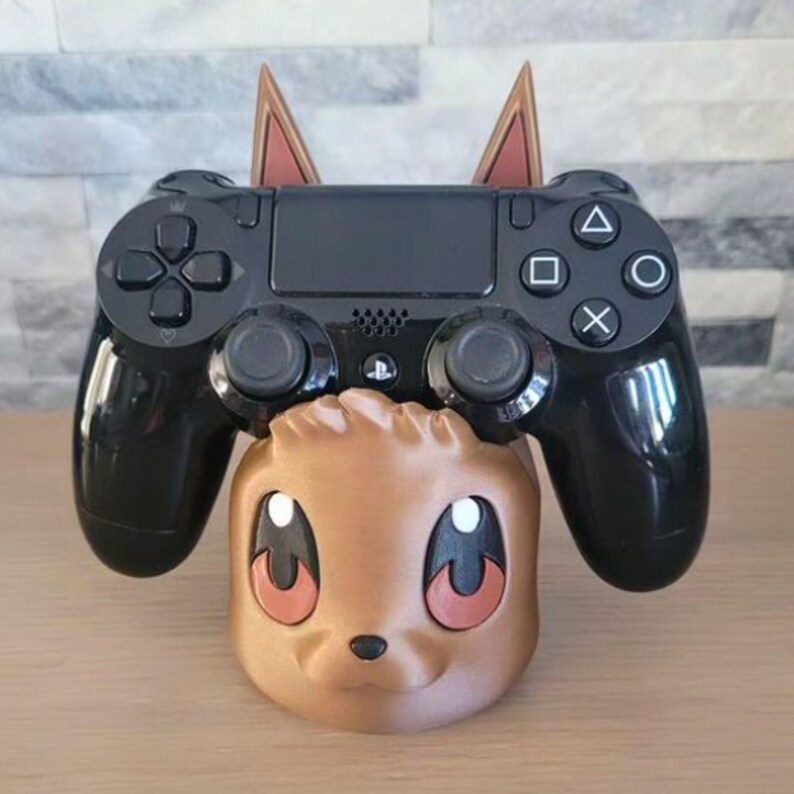 May include: A black PlayStation 4 controller rests on a brown and tan character-themed stand. The stand has large eyes and ears. The controller has the standard button layout.