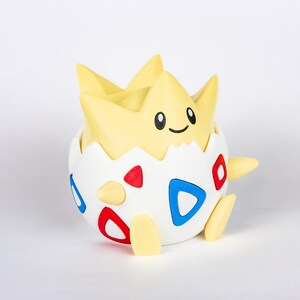 Puede incluir: Figura de Togepi, un personaje de Pok&eacute;mon. La figura es principalmente blanca con detalles rojos y azules. La parte superior de la figura es amarilla con una cara sonriente. La figura tiene forma de huevo.