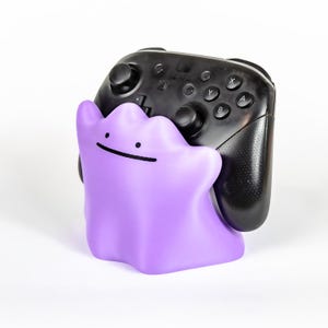 May include: A purple Ditto-shaped controller holder with a black Nintendo Switch controller. The holder has a simple face design. The controller is black with buttons labeled A, B, X, and Y.