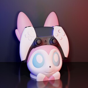 May include: A pink and white character-themed controller holder with a PlayStation 5 controller. The holder has large pink ears and a smiling face with blue eyes. The controller is black and white.