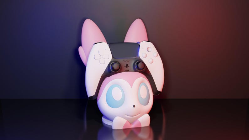 May include: A pink and white character-themed controller holder with a PlayStation 5 controller. The holder has large pink ears and a smiling face with blue eyes. The controller is black and white.
