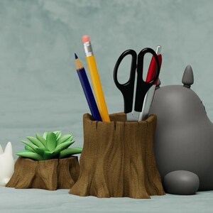 May include: A gray Totoro character figurine, a white Totoro figurine, and a brown wooden tree stump pencil holder with a green succulent plant. The pencil holder has two pencils and a pair of scissors inside.