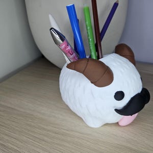 May include: White ceramic desk organizer shaped like a cartoon animal with brown horns, black mustache, and pink tongue. The organizer has multiple pens and pencils inside.