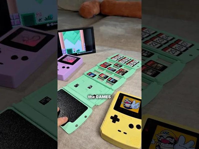 May include: A collection of retro handheld gaming consoles and game cartridges. The image features a yellow and a purple console, along with a green cartridge holder. The text "the GAMES" is visible on the green holder.