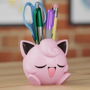 Puede incluir: Un organizador de escritorio Pokemon Jigglypuff rosa y negro con una cara sonriente. El organizador sostiene un par de tijeras azules, un bol&iacute;grafo verde, un bol&iacute;grafo morado y un l&aacute;piz amarillo.