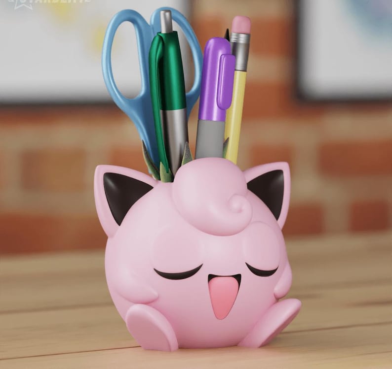 Puede incluir: Un organizador de escritorio Pokemon Jigglypuff rosa y negro con una cara sonriente. El organizador sostiene un par de tijeras azules, un bol&iacute;grafo verde, un bol&iacute;grafo morado y un l&aacute;piz amarillo.