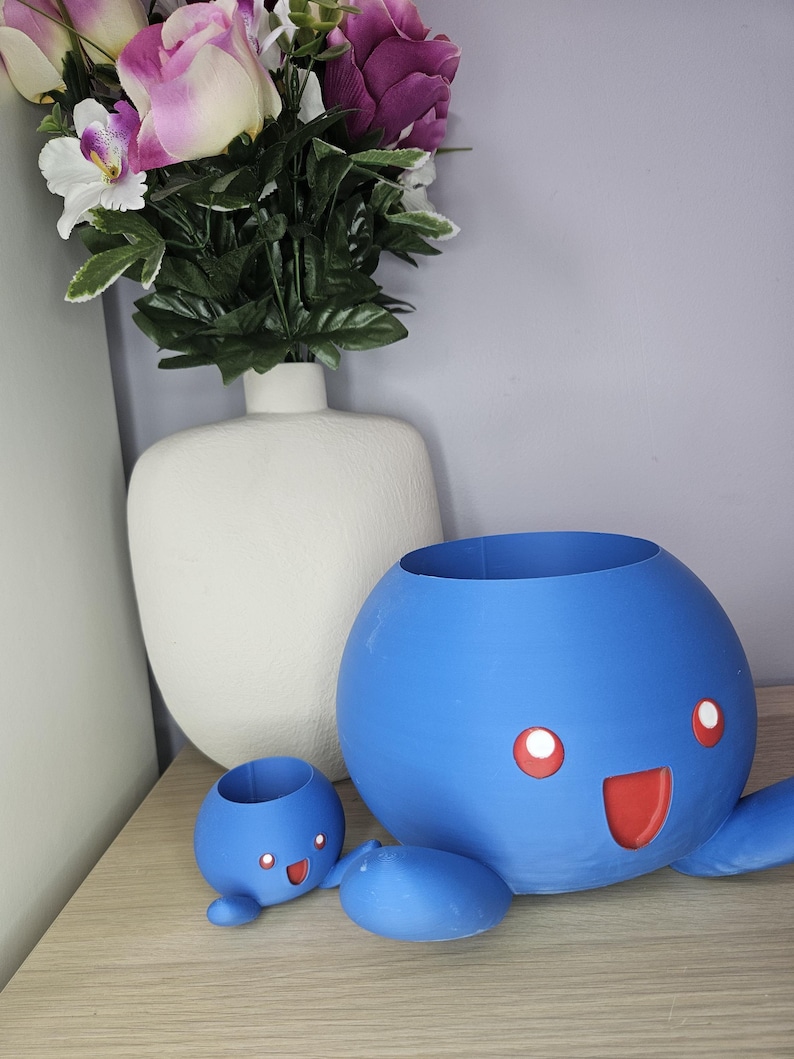 May include: Two blue 3D printed planters shaped like a cartoon character with a smiling face and red eyes. The larger planter has two arms and a lid. The smaller planter has two arms and is sitting on a wooden surface.