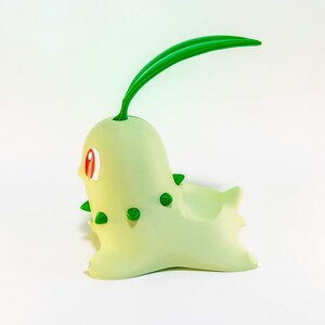 May include: A light green Chikorita figurine with a bulbous body, green spikes, and a long green leaf on its head. The figurine has red eyes and is set against a white background.