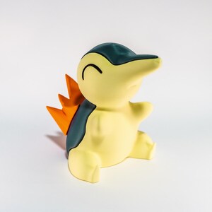 May include: A Cyndaquil figurine, a Pok&eacute;mon character, is shown in a cheerful pose. The figure is mainly yellow with a dark teal back and head, and orange flames on its back. The figurine is sitting down.