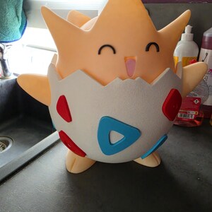 May include: A light orange Togepi figure, with a smiling face and small black eyes. The figure is emerging from a white eggshell with red and blue accents. The figure has outstretched arms and is standing on a dark surface.