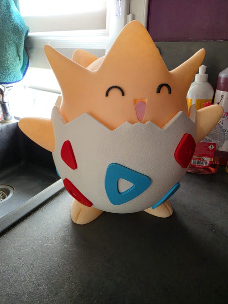 May include: A light orange Togepi figure, with a smiling face and small black eyes. The figure is emerging from a white eggshell with red and blue accents. The figure has outstretched arms and is standing on a dark surface.